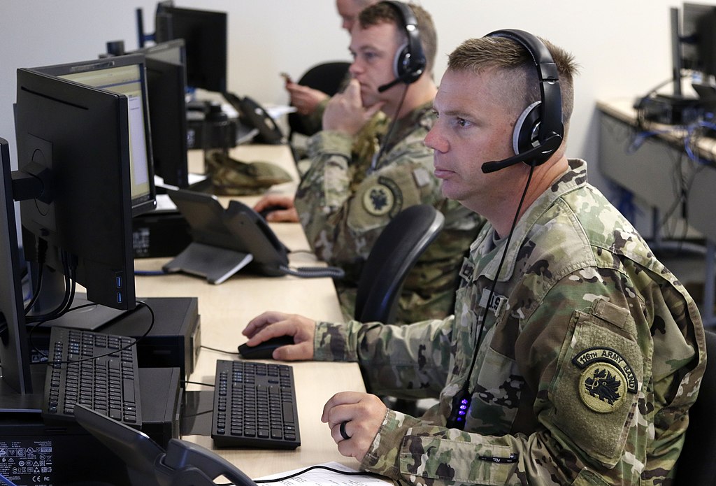 Call center of the Georgia National Guard with white men at computers wearing camo fatigues and headsets, one man looking at phone rather than at computer monitor. One soldier, in the middle of the frame has his thumb under his chin and finger on his lip.